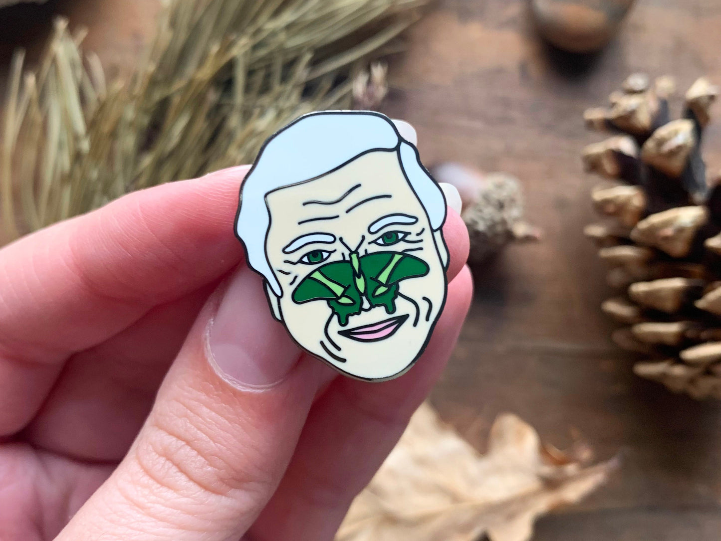 Enamel pin of Sir David Attenborough with a green butterfly on his face.