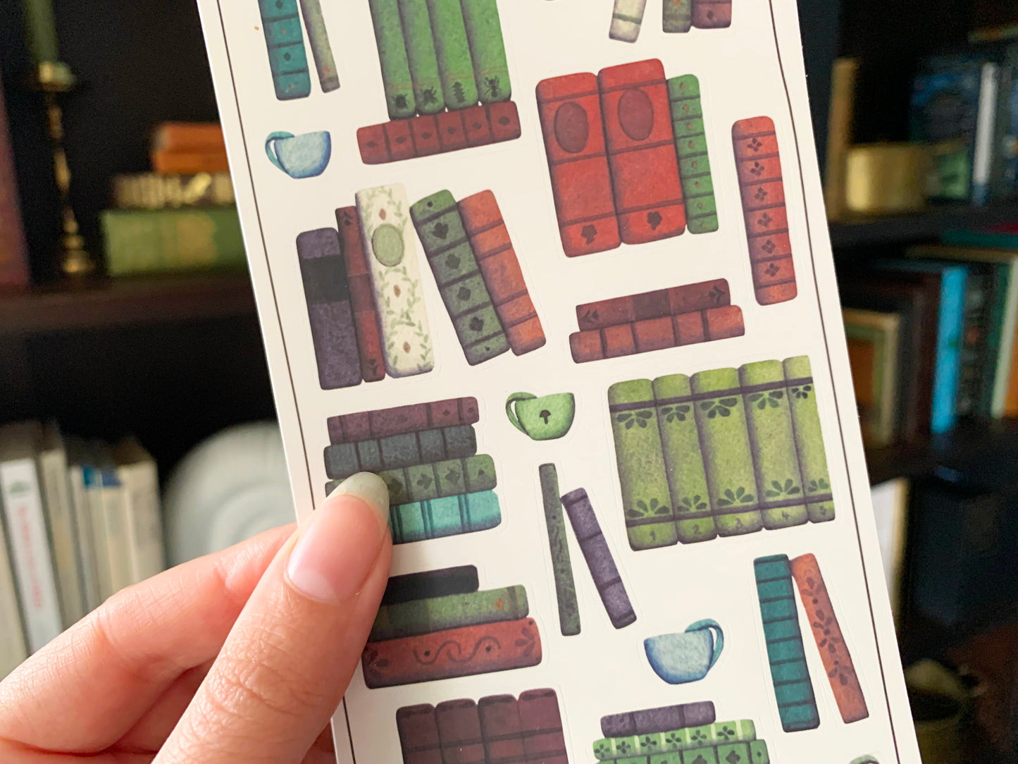 Close up of a sticker sheet of a vintage books and cups of tea to be used in planner or journal, held in front of a dark academia style bookshelf. The Naturalist's Guild.